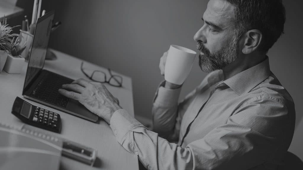 Mann mit Bart sitzt im HomeOffice am Tisch, vor ihm steht ein geöffnetes Laptop, ein Taschenrechner und eine Brille. Er führt mit der rechten Hand eine Tasse zum Mund.