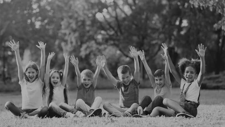 Lachende Kinder sitzen im Freien auf dem Rasen und halten ihre Hände in die Luft.