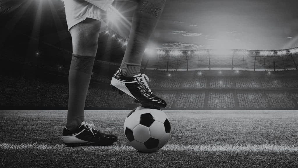 Ausschnitt eines Fußballstadions ist im Hintergrund des Bildes, Beine eines Fußballers, der über seine Unternehmensziele nachdenkt, sind zu sehen. Das rechte Bein steht auf einem Ball.